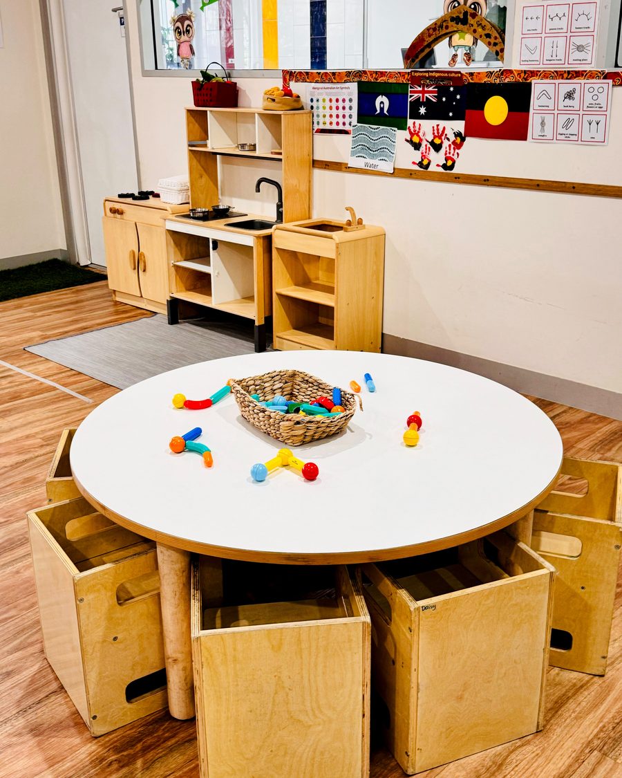 A round table with building pieces, cube seats and a wooden play kitchen in a Blooming Buds room